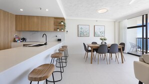 A modern kitchen with a central island, bar stools, a dining table with chairs, and a view of the outdoors through large windows.