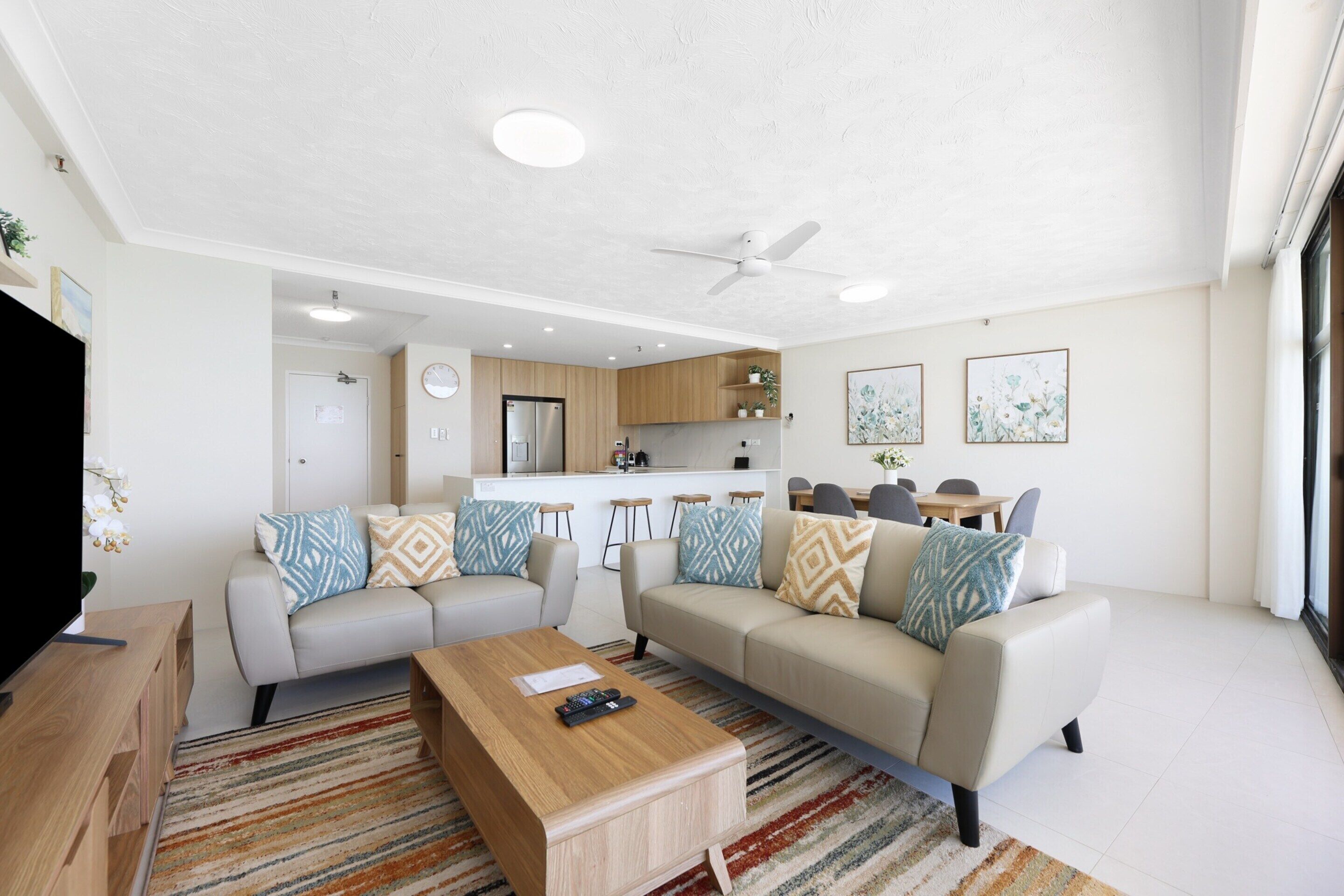 A modern living room with a sofa, coffee table, and a TV. There is a kitchen area with wooden cabinets and a refrigerator.