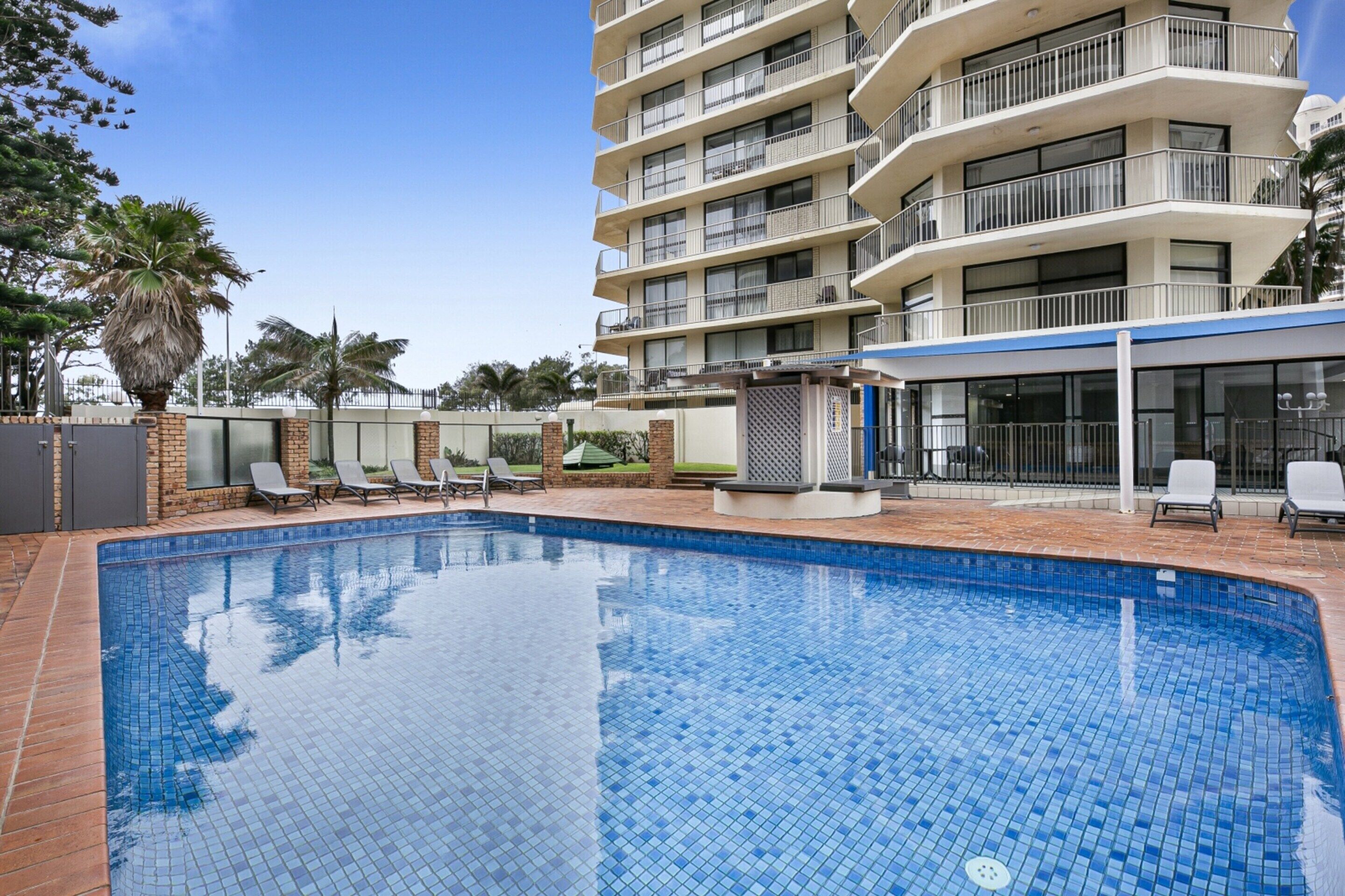 A swimming pool with tiled edges, surrounded by lounge chairs and a multi-story building with balconies.