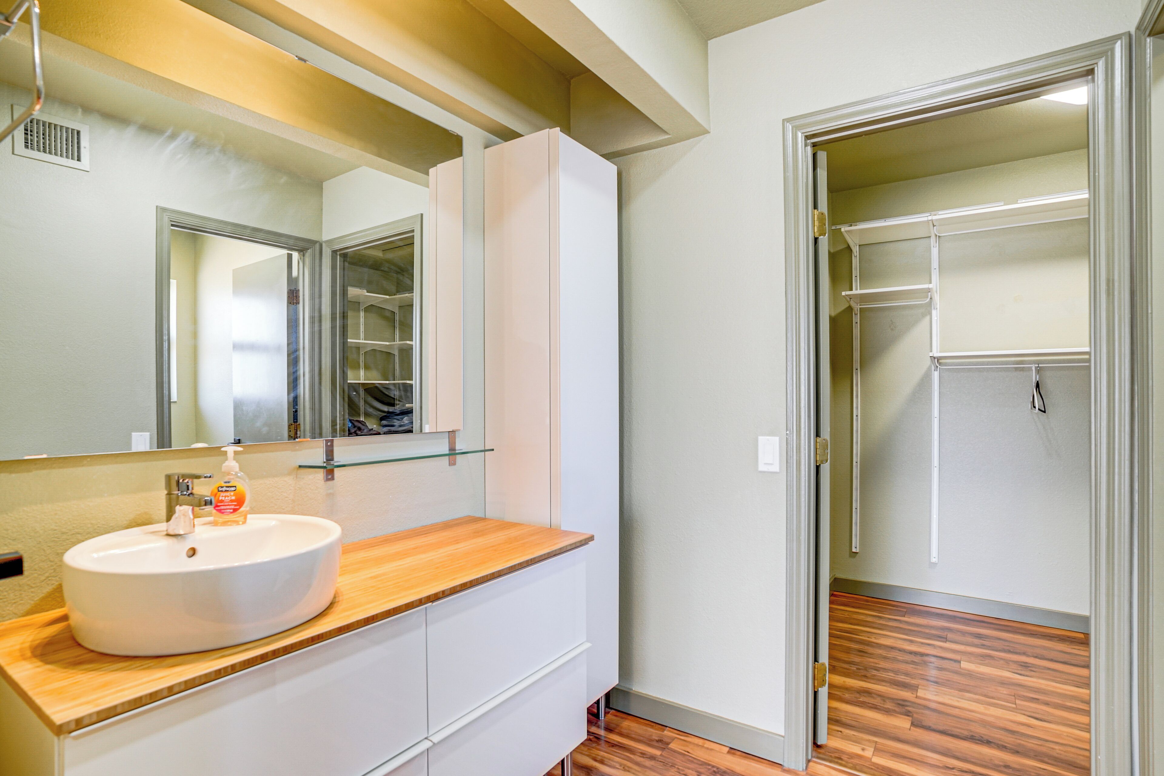 A bathroom with a white sink, wooden countertop, and a mirror above it. There is a white cabinet below the countertop and a wooden floor.
