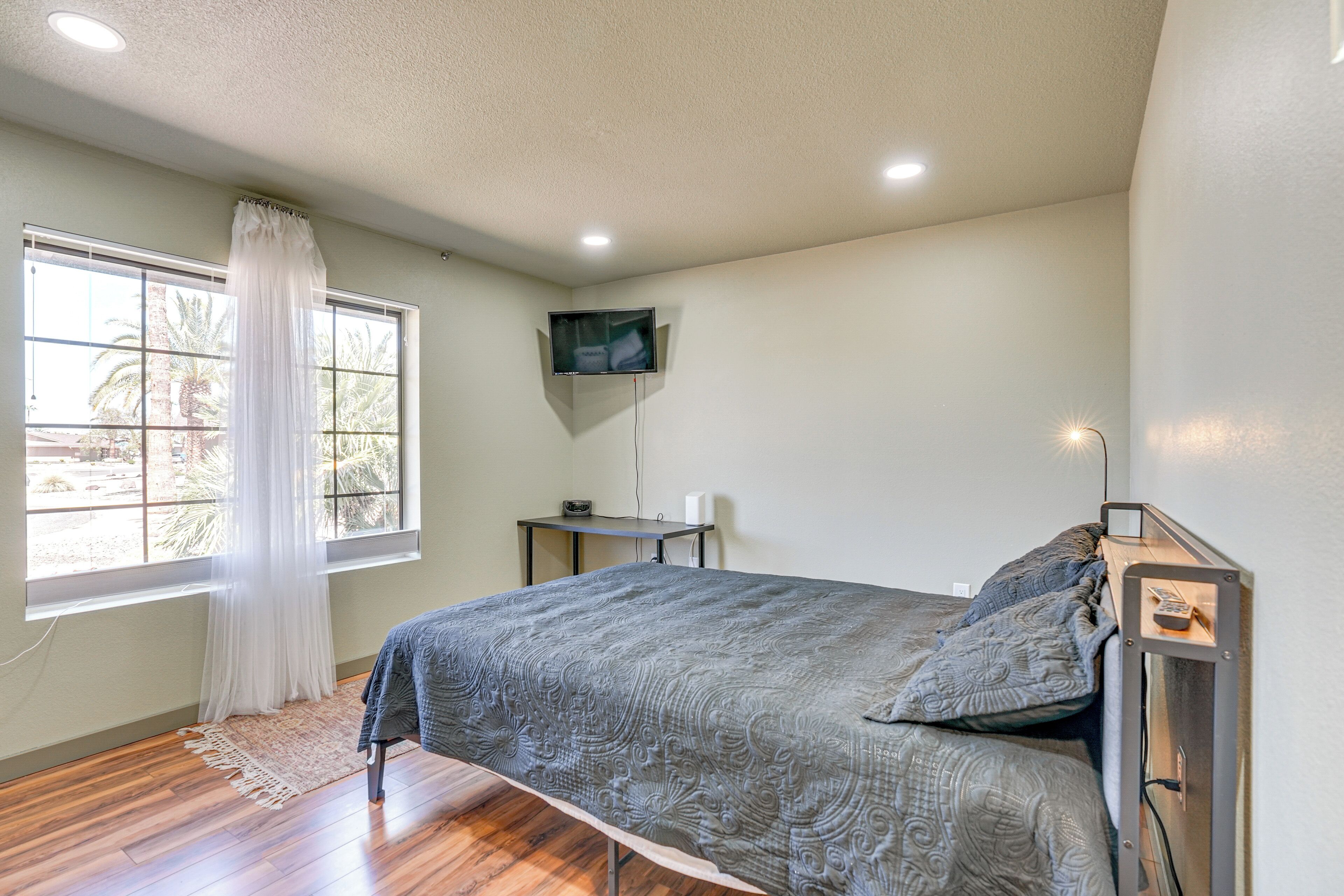 A bedroom with a bed, a TV mounted on the wall, a desk, and a window with curtains.