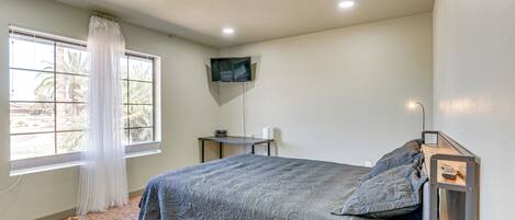 A bedroom with a bed, a TV mounted on the wall, a desk, and a window with curtains.