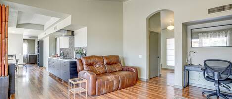 A spacious living area with a leather sofa, wooden flooring, a small side table, and a desk with a chair.