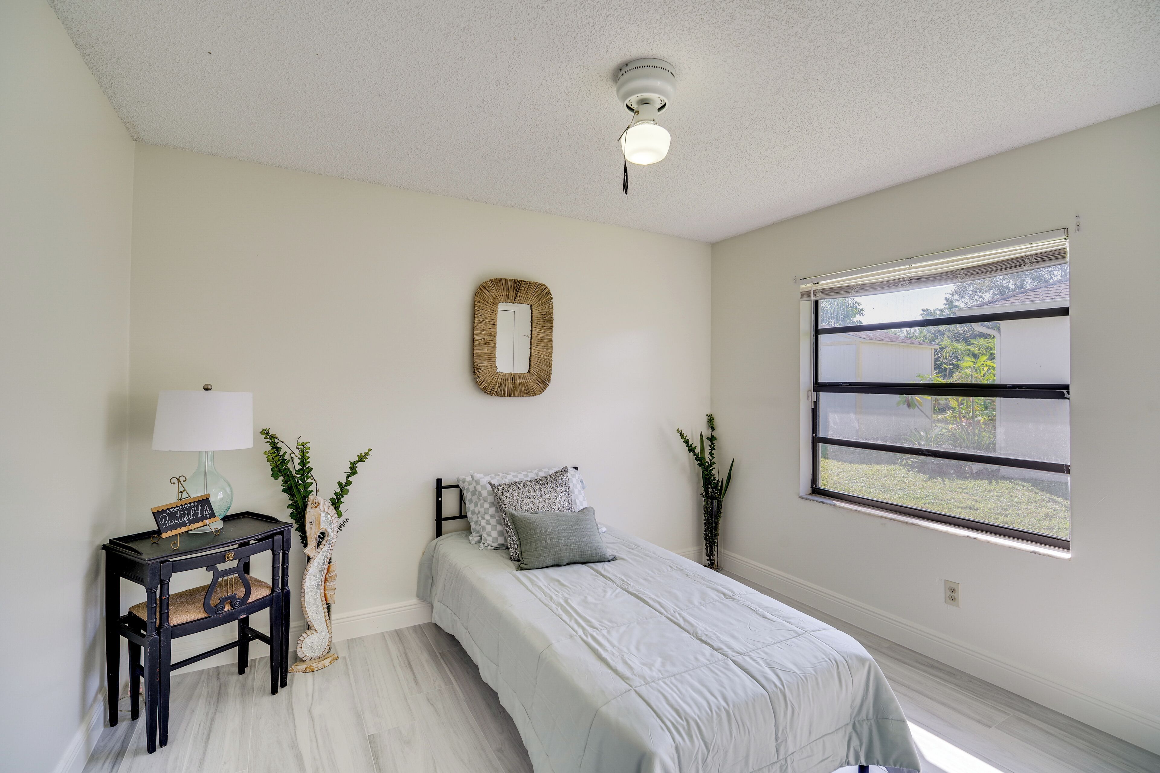 A bedroom with a bed, a bedside table with a lamp, a mirror, and a window with a view of greenery.