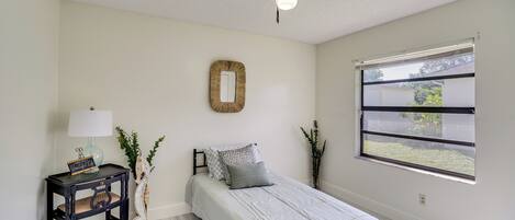 A bedroom with a bed, a bedside table with a lamp, a mirror, and a window with a view of greenery.