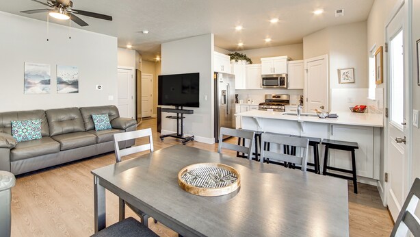Una cocina moderna con armarios blancos, microondas y refrigerador. Hay una zona de comedor con una mesa y sillas, y un sofá gris con cojines estampados.