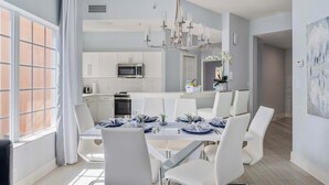 A modern dining area with a white table set for six, a kitchen in the background, and a large window with curtains. - Serene Disney Getaway, Designer Style, MP61 (Orlando)