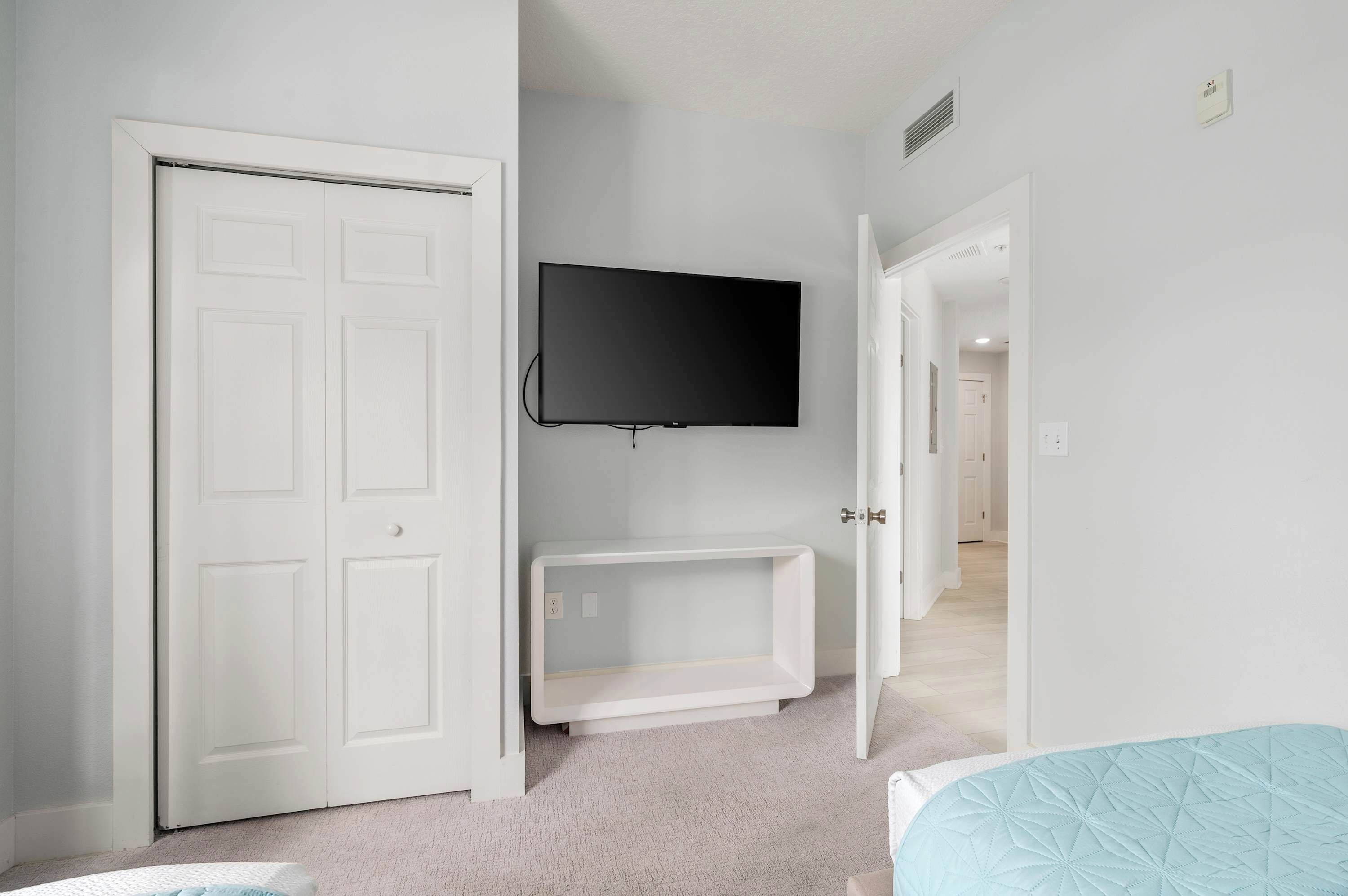 A room with a white door, a mounted TV, a white cabinet, and a bed with a blue quilt.