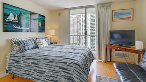 A hotel room with a large bed, a TV, a desk, and a view of a cityscape through the window. - Walkable Urban Retreat! Condo in Honolulu (Honolulu)