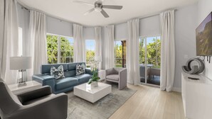 A modern living room with a blue sofa, a grey armchair, a white coffee table, a ceiling fan, and large windows with curtains. - Sleek & Stylish Stay, Mins to Magic, MP68 (Orlando)
