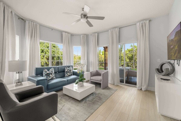 A modern living room with a blue sofa, a grey armchair, a white coffee table, a ceiling fan, and large windows with curtains.