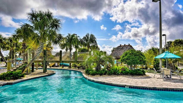 A clear blue pool with a wooden bridge, surrounded by palm trees and a thatched-roof structure. - Championsgate Resort Perks! Fun-filled Family Home (Davenport)