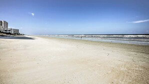 Ein Strand mit blauem Himmel, einigen Gebäuden im Hintergrund und einer Person, die am Ufer entlanggeht.