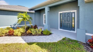 A modern house with a blue exterior, a front door, and a landscaped garden with various plants. - Fish, Golf & Beach-hop! Family Home in Cape Coral (Cape Coral)