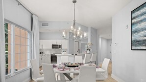 Une salle à manger moderne avec un lustre, des chaises blanches, une table à manger dressée et une vue sur la cuisine en arrière-plan.