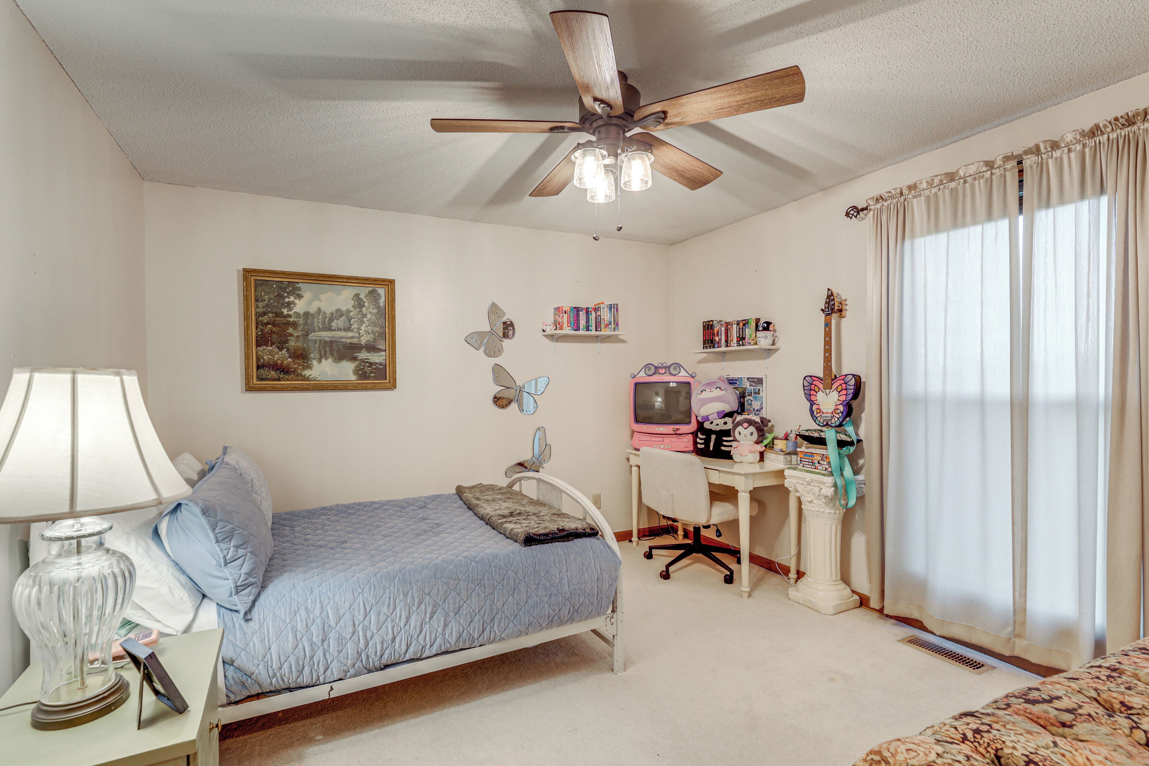 A bedroom with a bed, a desk, a chair, a ceiling fan, and a window with curtains.
