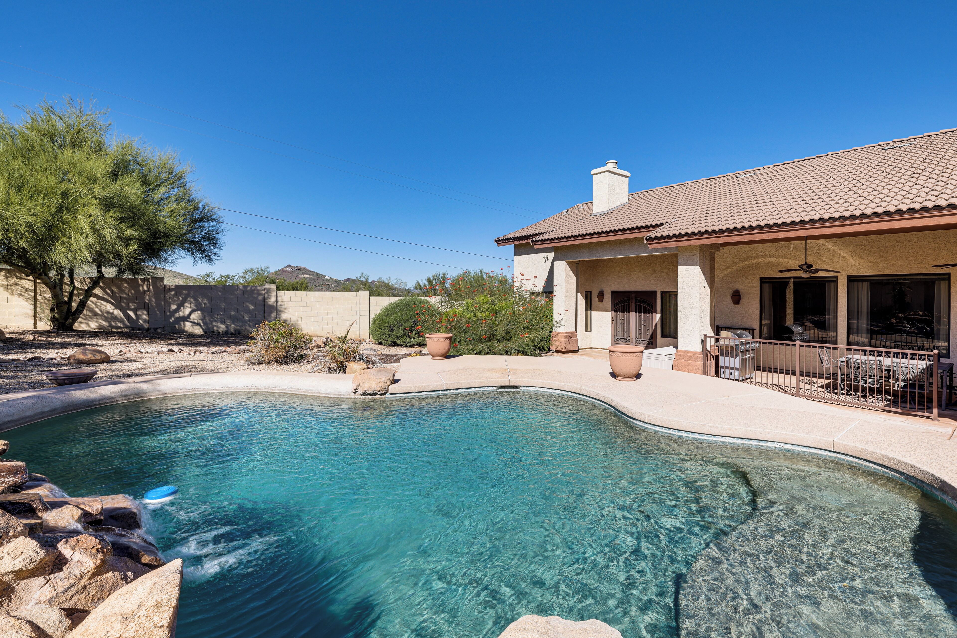 A clear swimming pool with a tiled deck, surrounded by a house with a tiled roof and a fenced patio area.