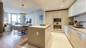 A modern kitchen with built-in appliances, a dining area with a round table and chairs, and a view of the city through large windows. - Luxe Condo w/ Hallandale Beach Oceanfront View! (Hollywood)