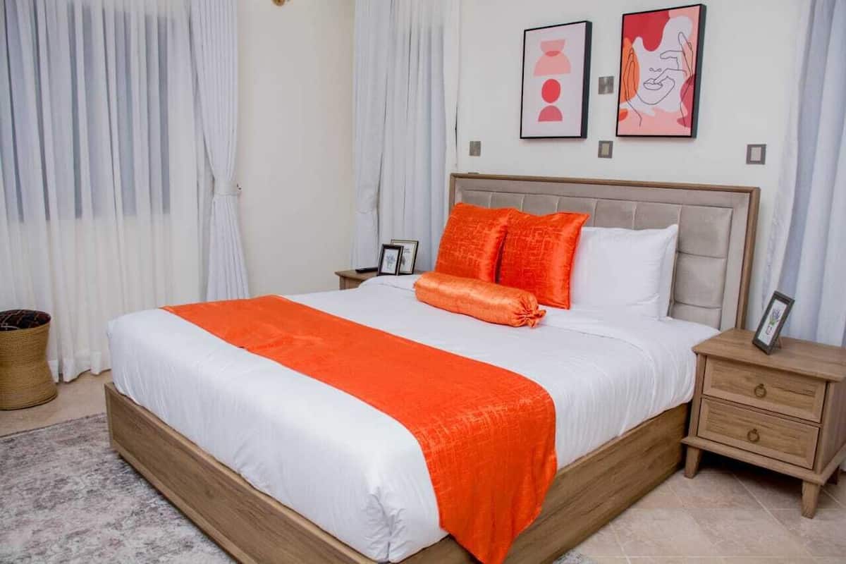 Uma cama bem arrumada com colcha branca e laranja, um painel de cabeceira de madeira e duas mesas de cabeceira com quadros.