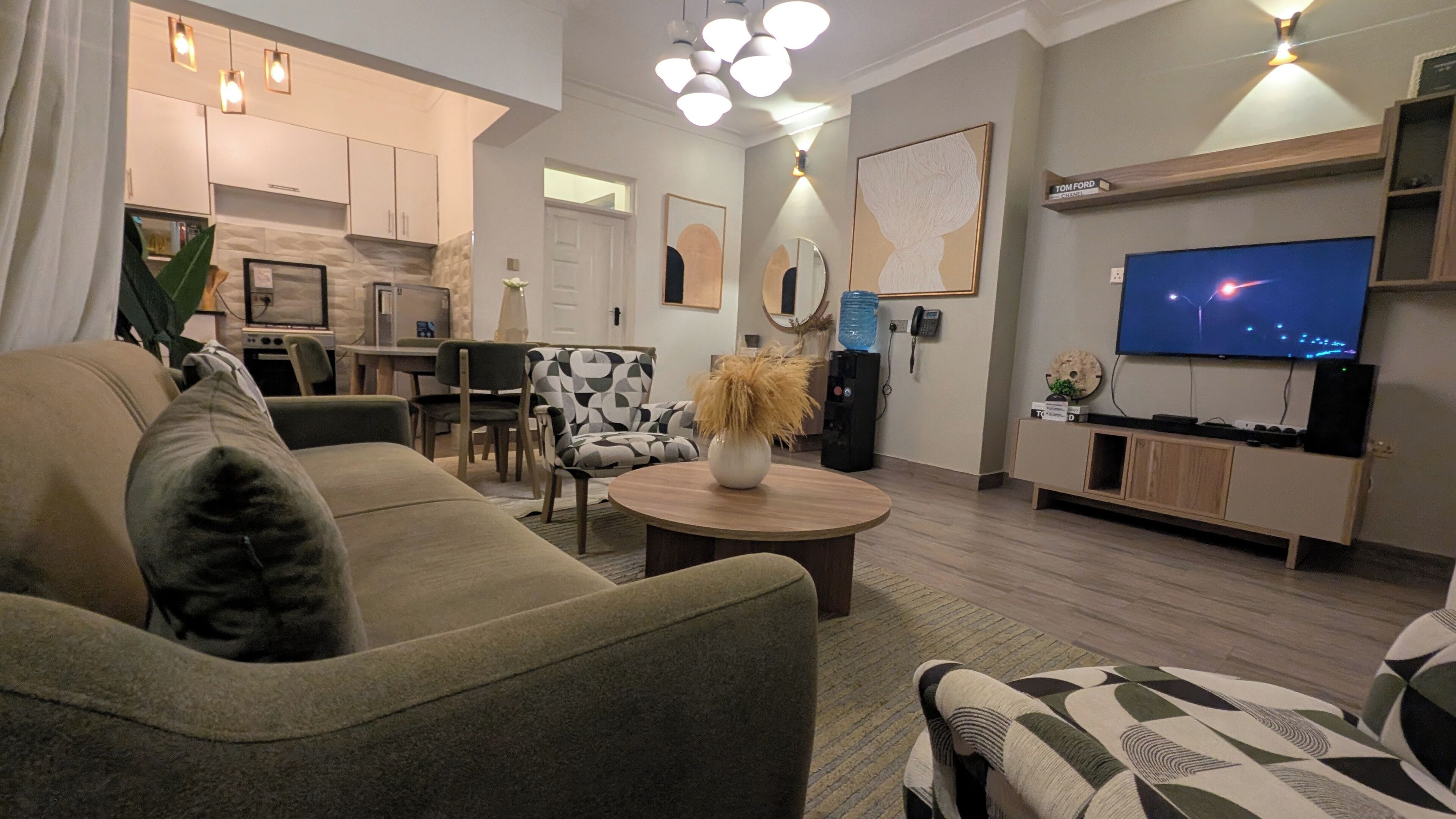 A modern living room with a sofa, coffee table, and a TV. There is a kitchen in the background with cabinets and a water dispenser.