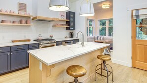 A modern kitchen with a central island, bar stools, pendant lights, and wooden shelves. - 2 Mi to Payette Lake: Charming Cottage w/ Hot Tub! (McCall)