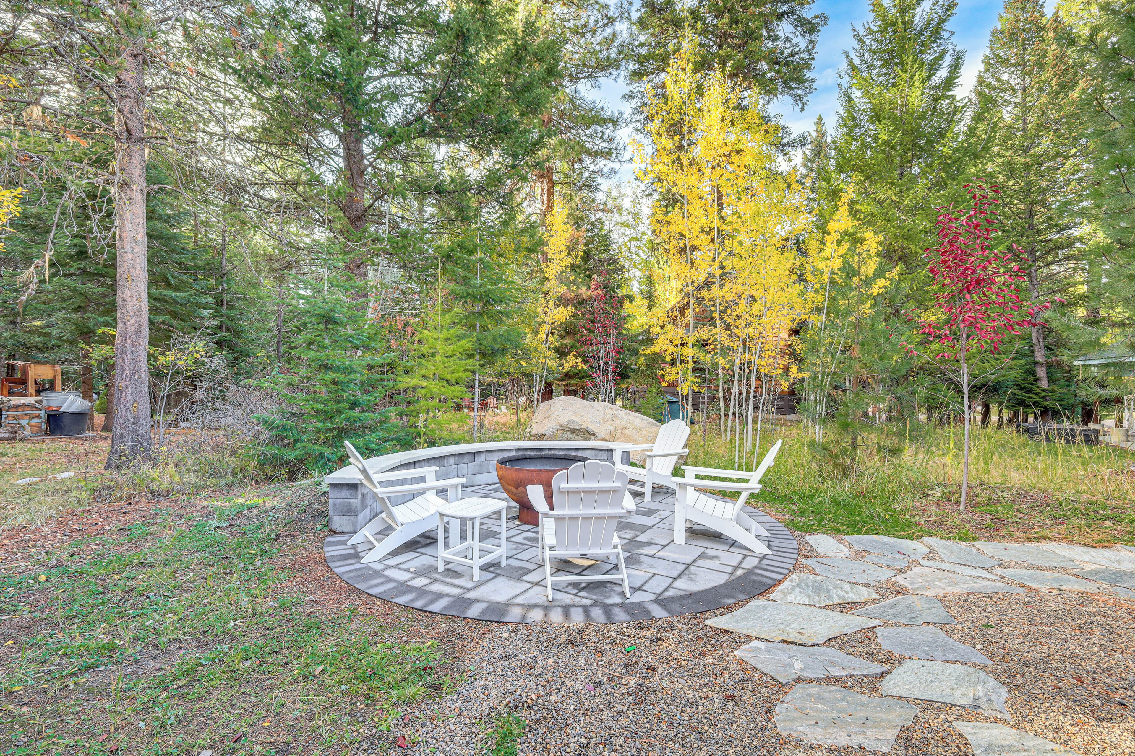 A circular outdoor seating area with stone flooring and white Adirondack chairs, surrounded by a forest with autumn-colored trees.