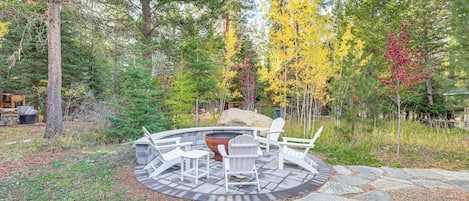 A circular outdoor seating area with stone flooring and white Adirondack chairs, surrounded by a forest with autumn-colored trees.