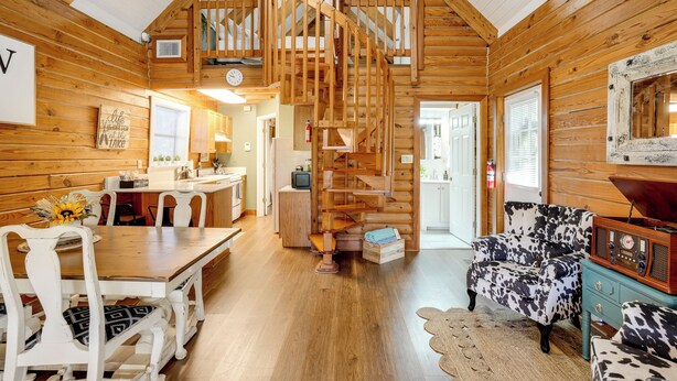 A log cabin interior with a wooden staircase, a dining area with white chairs, a cow print sofa, and a vintage radio. - Lake Okeechobee Dockside Cabin w/ Pool Access (Okeechobee)