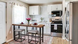 Una cocina con armarios blancos, una mesa de madera con dos taburetes, un refrigerador de acero inoxidable y un microondas.