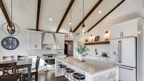 A modern kitchen with white cabinetry, a large island with bar stools, wooden beams on the ceiling, and pendant lighting. - Mtn Views, Hot Tub + Fire Pit: Black Hills Retreat (Piedmont)
