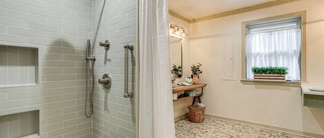 A bathroom with a shower, a sink, a window with a plant, and a tiled floor.