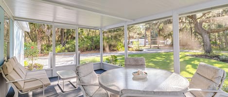 Une terrasse couverte avec une table ronde en verre et quatre chaises rembourrées, entourée de grandes fenêtres offrant une vue sur une pelouse bien entretenue et des arbres.
