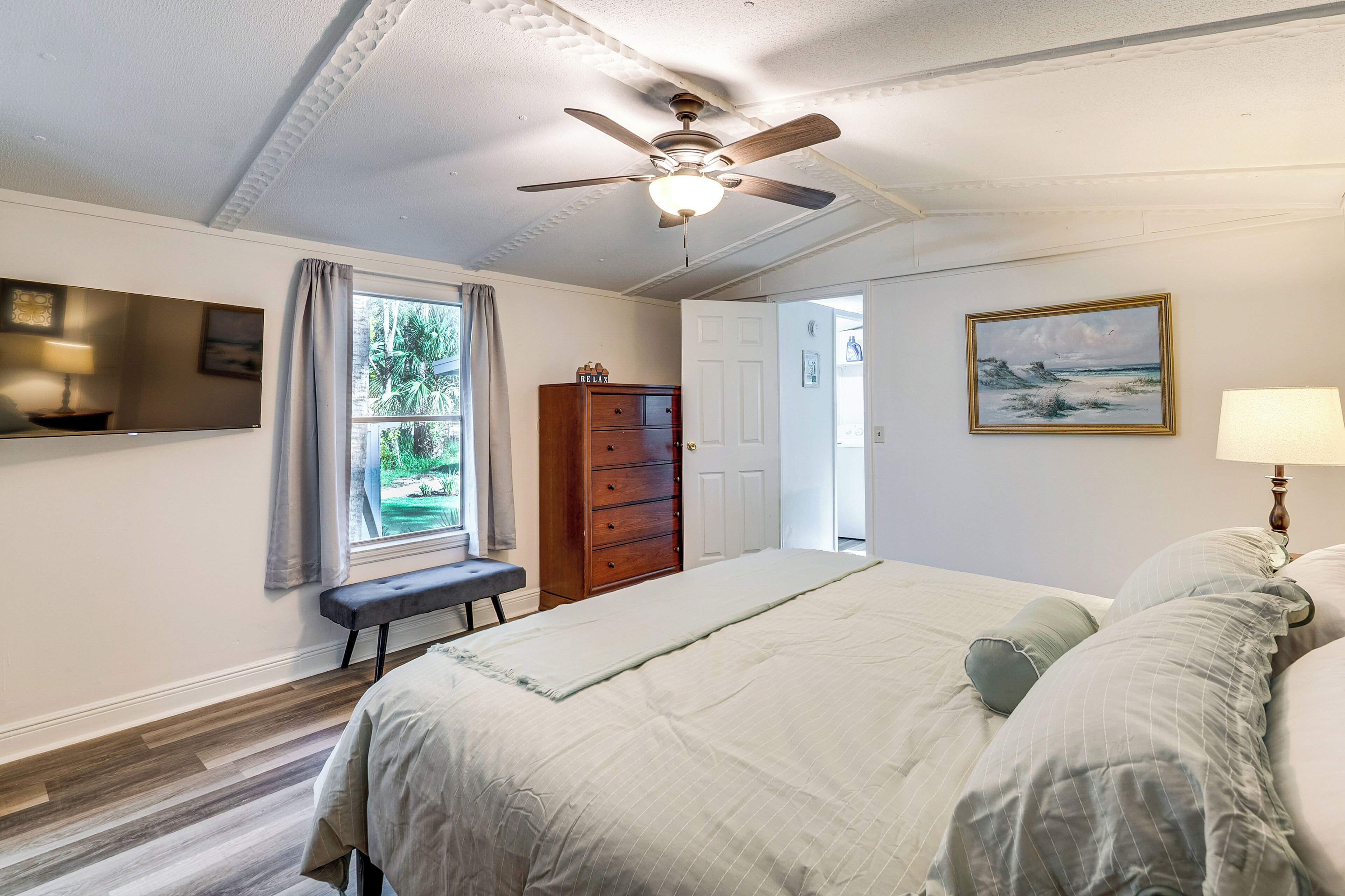 A bedroom with a bed, a ceiling fan, a wooden dresser, a bench, and a window with curtains.