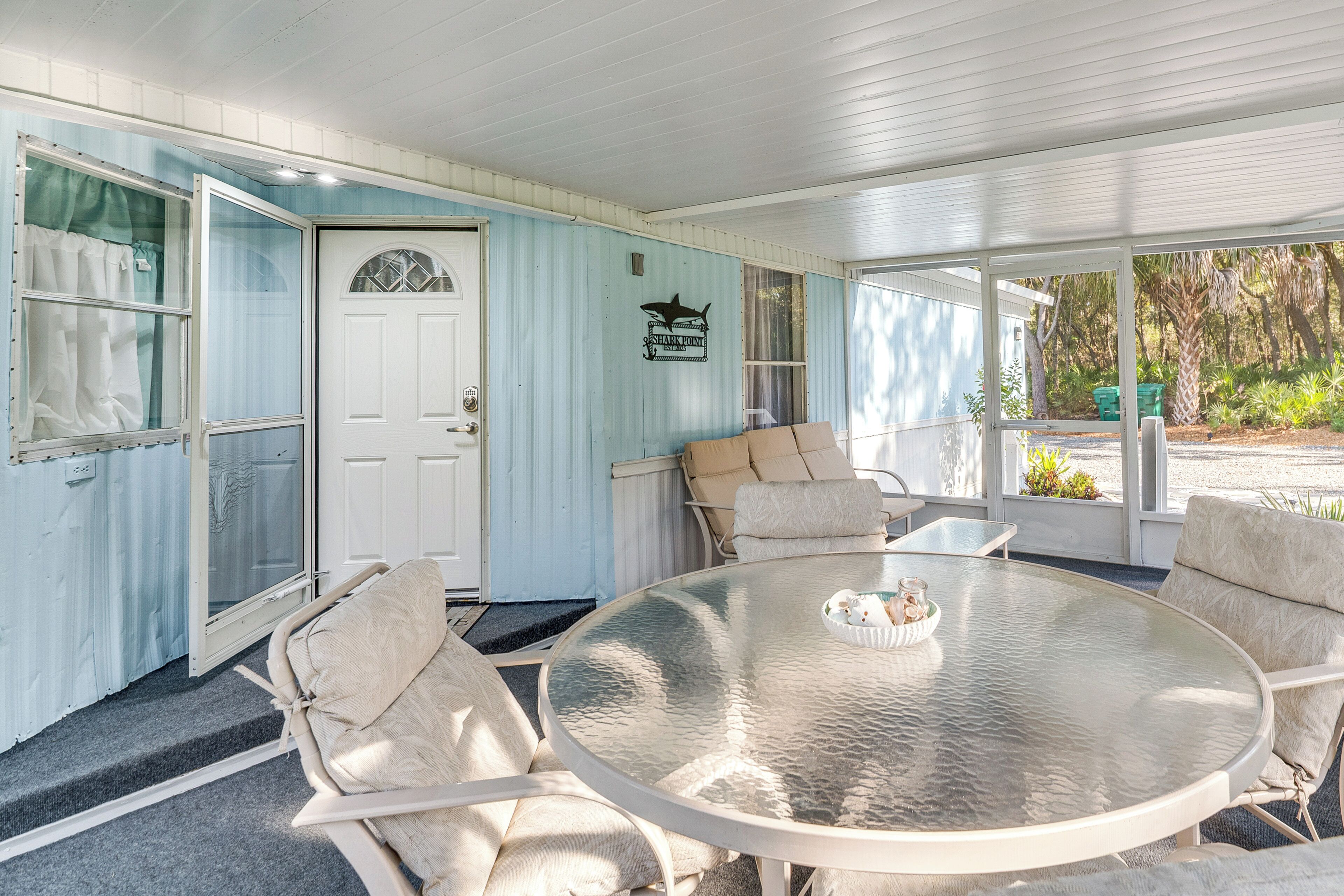 A covered porch with a round glass table, cushioned chairs, and a door with a decorative glass panel.
