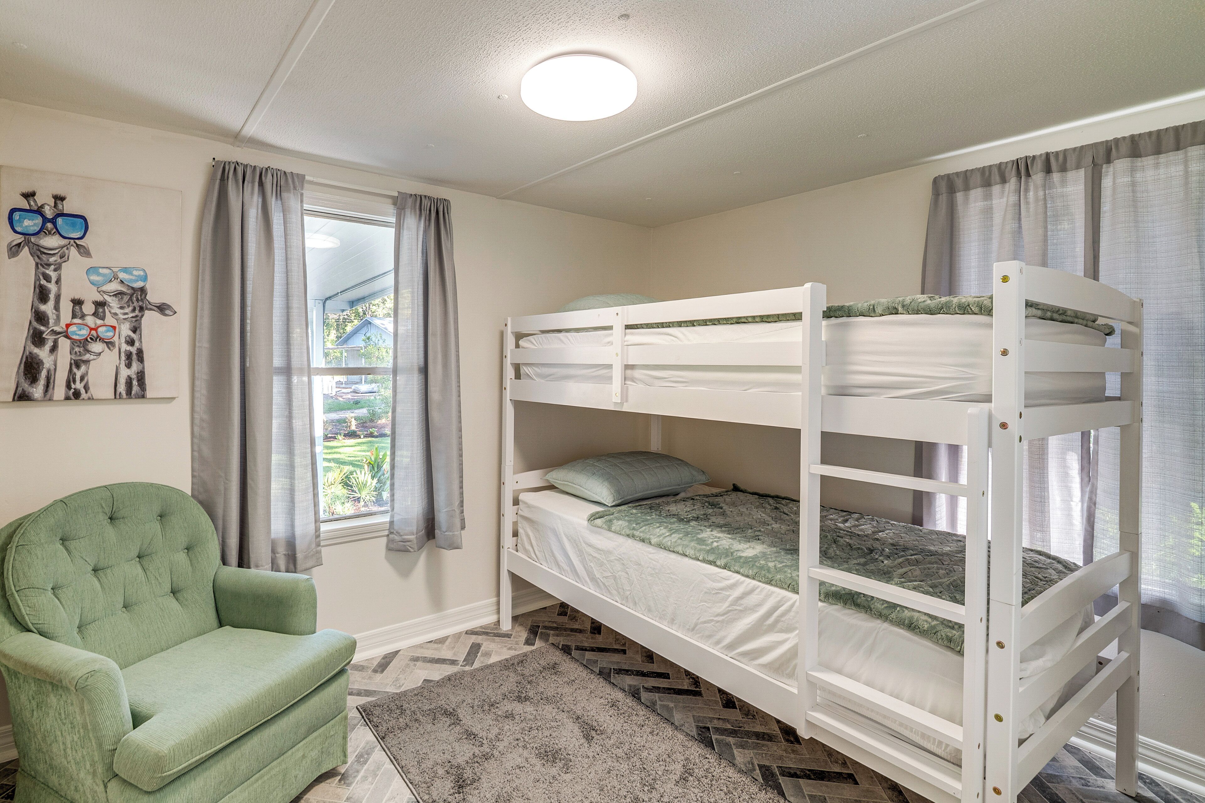 Habitación con literas, un sillón verde, un cuadro de jirafas y una ventana con cortinas.