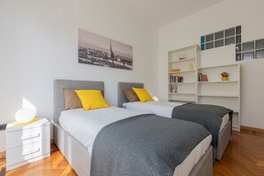 Ett modernt sovrum med två sängar, ett nattduksbord, en bokhylla och en tavla med en stadsbild på väggen.