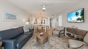 En moderne stue med en mørk lædersofa, et rundt sofabord og et spiseområde med træstole. I baggrunden ses et køkken med hvide skabe og en stor køkkenø.