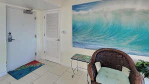 A room with a large wave painting on the wall, a wicker chair, and a small table with a decorative item. - Regency Towers #707 West | Sunset Views, Pool, Tennis/Pickleball (Pensacola Beach)