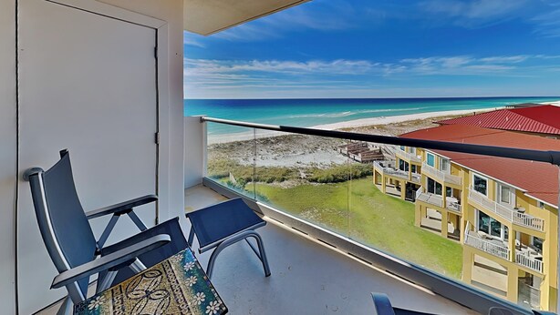 A balcony with a view of a beach and a building with a red roof. - Regency Towers #707 West | Sunset Views, Pool, Tennis/Pickleball (Pensacola Beach)
