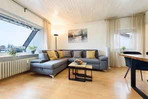 Una sala de estar moderna con un sofá seccional, una mesita con fruta, una lámpara de pie y vistas de la ciudad a través de amplios ventanales.