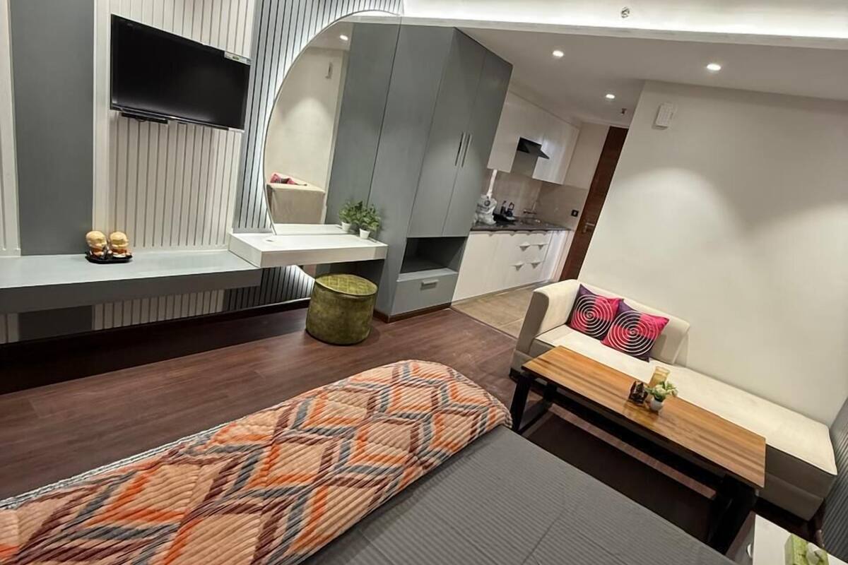 A modern hotel room with a bed, a sofa, a small table, a TV mounted on the wall, and a kitchen area in the background.