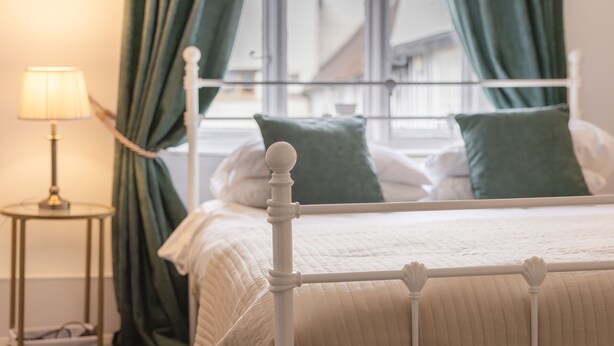 Un lit avec un cadre blanc et des oreillers verts, une lampe de chevet et une vue sur un immeuble à travers une fenêtre ornée de rideaux verts.