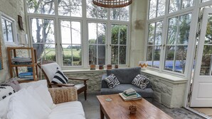 A sunroom with a white sofa, a grey sofa with patterned cushions, a wooden coffee table, and a bookshelf with books.