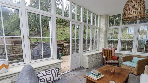 A sunlit room with large windows, a stone wall, a wooden table with books, and wicker furniture.