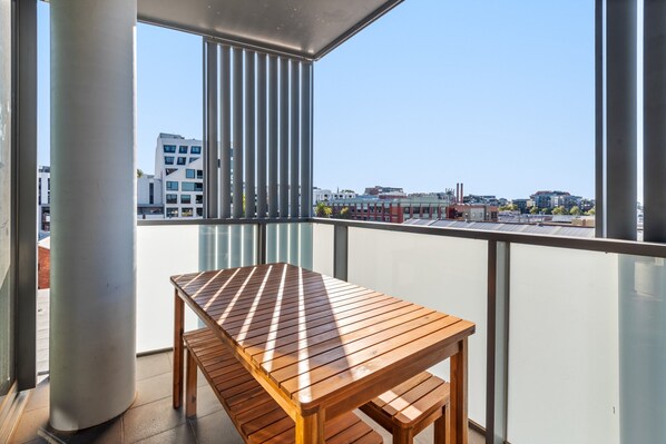 Un balcon avec une table en bois et des bancs, offrant une vue sur les bâtiments et un ciel dégagé.