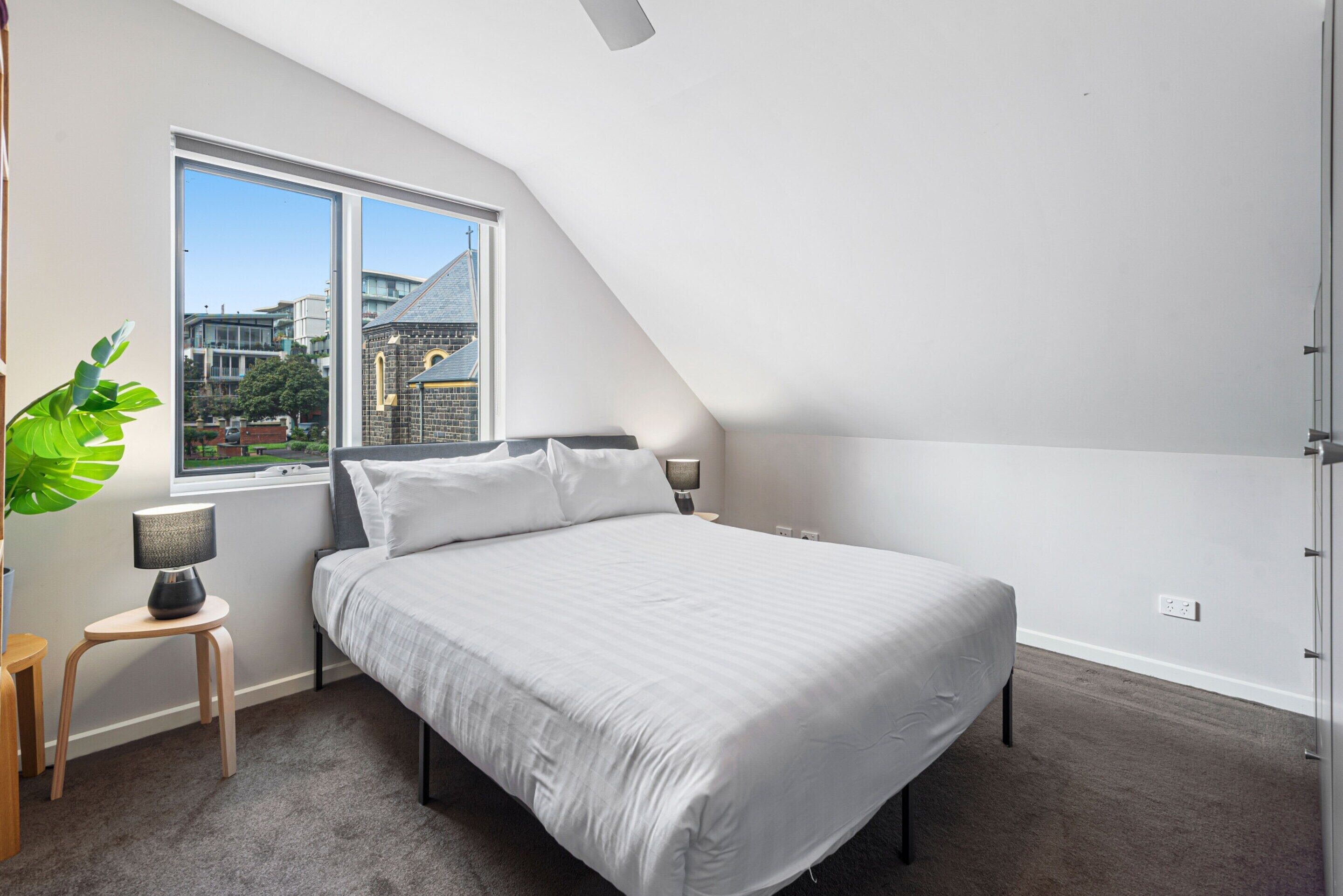 A modern bedroom with a large bed, a window with a view of buildings, a small table with a lamp, and a plant.