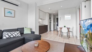 A modern living room with a sofa, a coffee table, and a TV. There is a kitchen area in the background with cabinets and a refrigerator. - Central 1-bed Apartment With Secure Parking (Melbourne)
