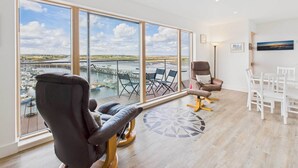 Une pièce spacieuse dotée de grandes fenêtres offrant une vue sur un port de plaisance et des bateaux, un coin salon confortable avec un fauteuil massant, une table à manger avec des chaises et un tapis à motifs sur le sol.