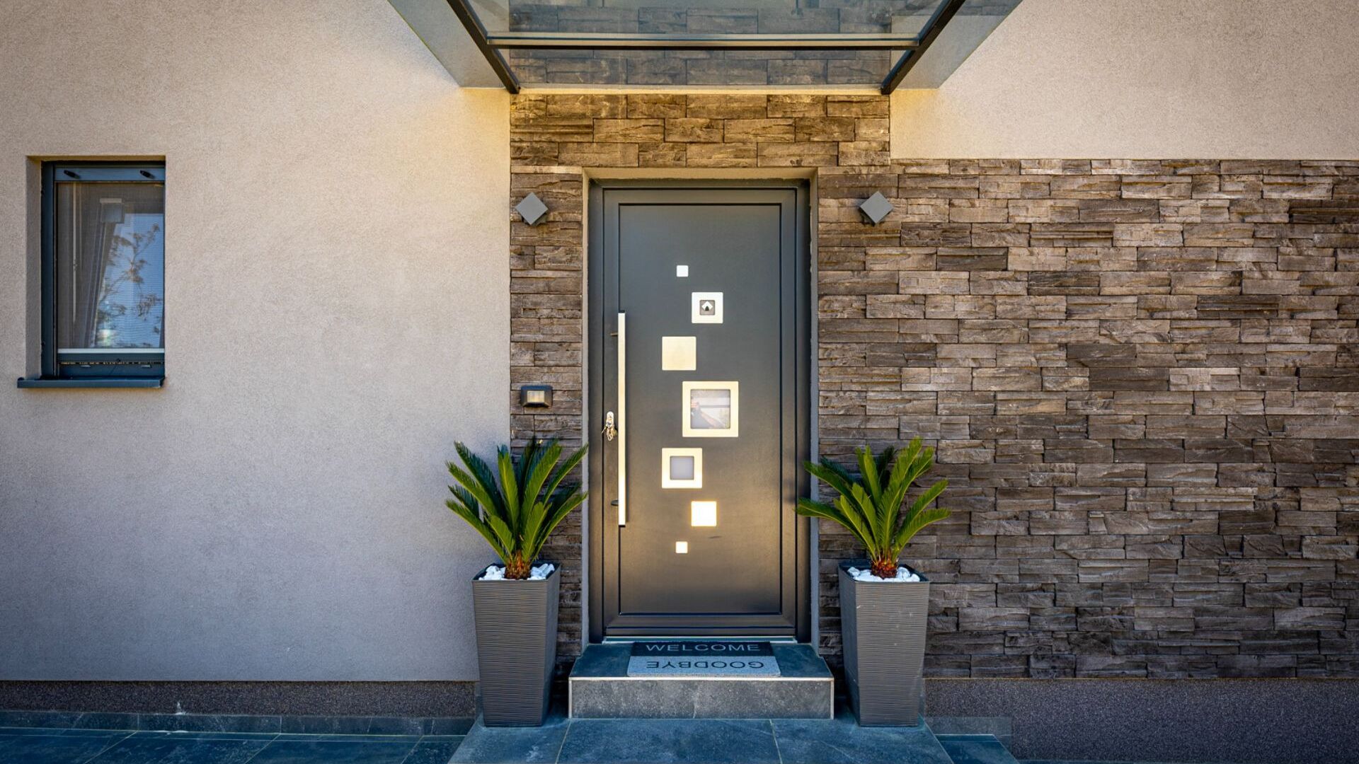 A modern building entrance with a stone facade, a dark door with illuminated panels, and two potted plants.