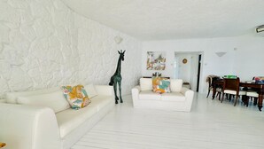 Un salon avec un mur blanc à texture, un canapé blanc, un fauteuil blanc plus petit et une statue de girafe. On y trouve également un coin repas avec une table et des chaises en bois.